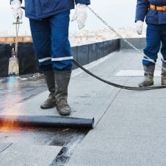 Two workers in protective clothing are installing roofing felt on a flat roof. One is using a blowtorch to heat and seal the material. They are standing on a city rooftop with safety harnesses and chains attached.