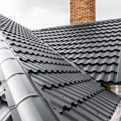 Close-up view of a house's roof with black metal roofing sheets. The roof features a steep slope design with a prominent red brick chimney. Partially cloudy sky and bare trees are visible in the background.