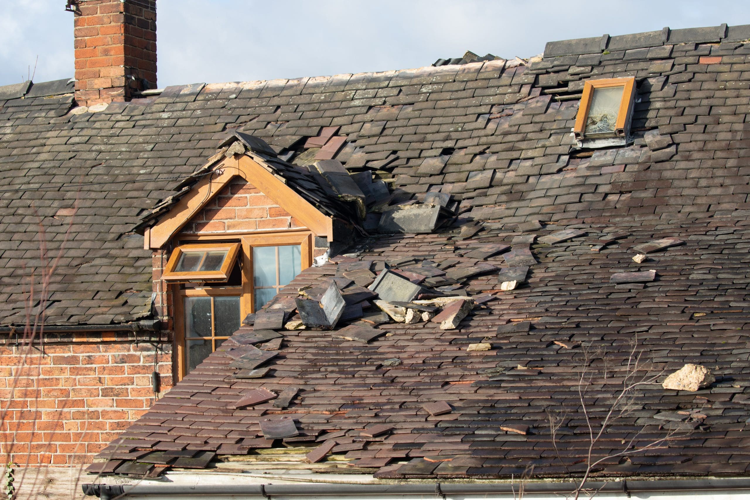 A damaged roof with missing and broken shingles. The roof has a dormer window and a skylight. Some shingles are scattered, and the underlying structure is visible, indicating the need for repair.