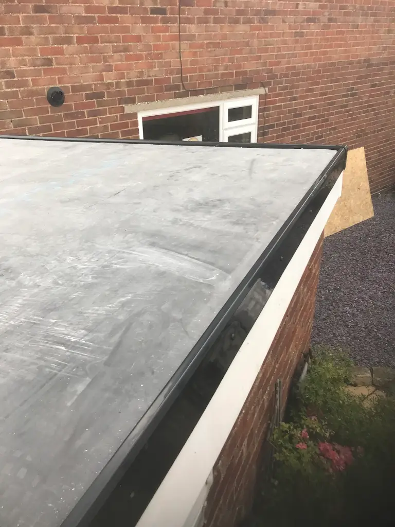 View of a flat roof with a rubber membrane surface, bordered by bricks, overlooking a garden and gravel path. A window and a partially visible wooden board are in the background against a brick wall.