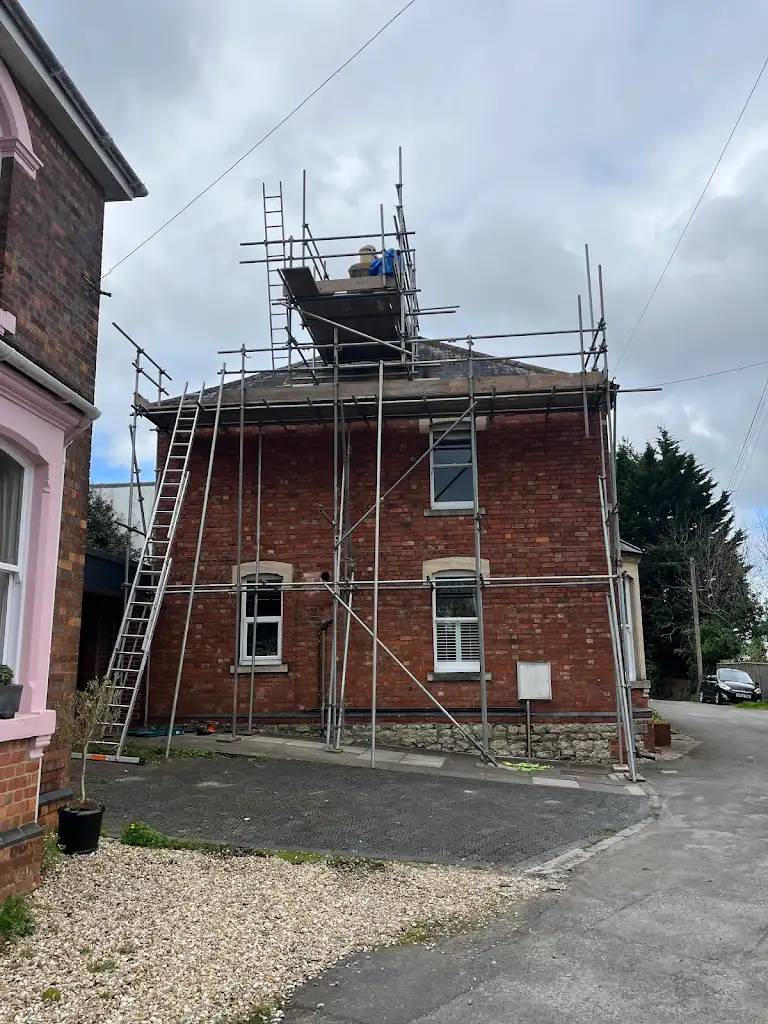A red brick house with scaffolding set up around the roof, indicating ongoing repairs or construction. The sky is cloudy, and there are two ladders leaning against the scaffolding. Another house is partially visible on the left.