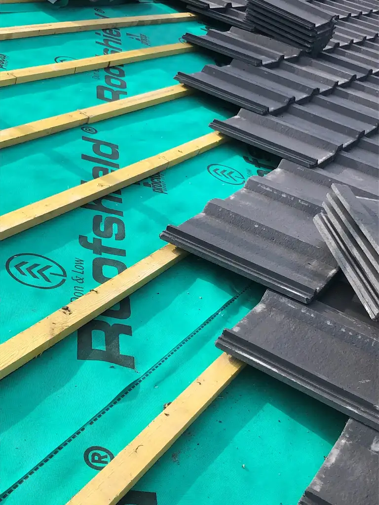 A roof under construction with wooden battens, green roof underlayment, and partially installed gray tiles. The text "Roofshield" is visible on the underlayment.
