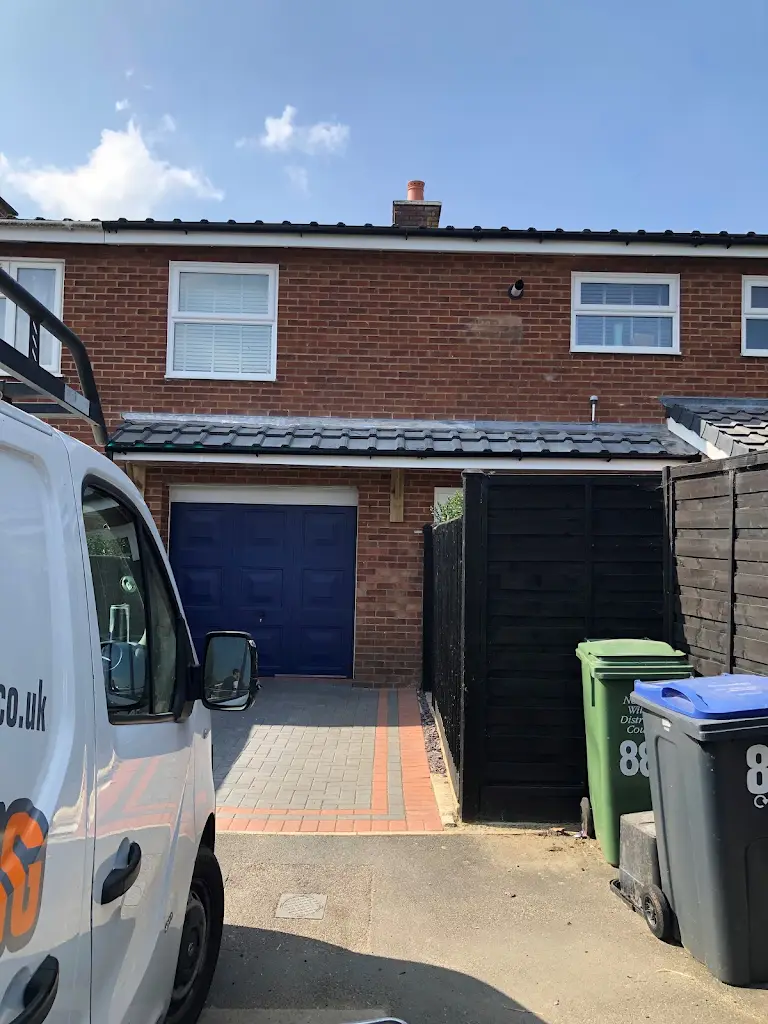 A two-story brick house with a blue garage door and two windows above. A white van is parked in the paved driveway. On the right, there are recycling and trash bins against a black fence under a clear blue sky.