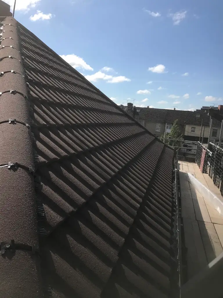 A rooftop lined with brown tiles overlooks a row of houses under a clear blue sky. A wooden platform is visible on the side, possibly part of a construction or renovation project.
